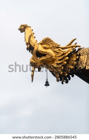 A sharp, detailed study of a gilded Makara gargoyle crowning the roof eave of a Tibetan Buddhist temple in Yunnan, China. The mythological sea creature is captured in stark profile against a pale, uniform sky, drawing immediate attention to the masterfully carved textures of its feathered gills, curling horn, and bared fangs. A delicate, weathered bronze wind bell hangs from its lower jaw, ready to catch the mountain breeze. The image exemplifies the transcendent metalwork and spiritual iconography that guards the sacred sanctuaries of the East.