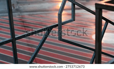 A sharp focus on angular metal bars creating a geometric frame over a blurred floor with red stripes and evening light reflections.