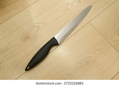 A sharp kitchen knife with a black handle resting on a wooden floor