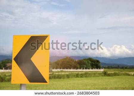 Sharp left curve warning chevron sign on a rural road against a sunny blue sky with clouds and a lush green field, symbolizing direction and journey.