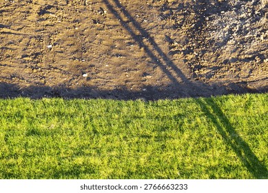Sharp linear division between green grass and dry soil with long shadows cast across surface in bright sunlight. Shows environmental contrast, land use separation, early landscaping stage, and visual geometry within urban outdoor space.