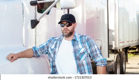 Sharp Looking American Truck Driver Wearing Blue Plaid Shirt And Black Baseball Cap With American Flag Stands Next To Big Rig. Casual Dressed Trucker On Job, Blue Collar Occupations In USA.