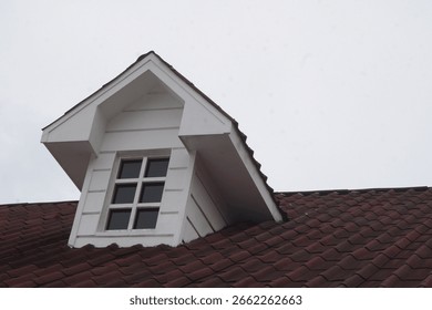 A sharp, modern dormer window with a clean white frame stands out against the textured red tiles of a classic pitched roof, set against a bright, overcast sky.