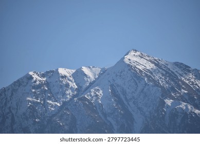 A sharp, pyramidal mountain peak rises prominently against a clear, solid blue sky. The steep slopes and jagged ridges of the summit are heavily blanketed with white snow and ice, emphasizing the deep vertical fissures and rocky contours of the mountain face.

To the left, additional craggy mountain peaks extend along the massive ridge line, also coated in patches of winter snow. Direct, bright daylight illuminates the right-facing slopes, casting the deep crevices and opposite aspects into crisp, dark shadows that contrast sharply with the sunlit snowy heights.