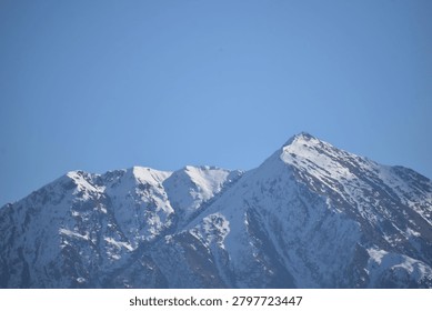 A sharp, pyramidal mountain peak rises prominently against a clear, solid blue sky. The steep slopes and jagged ridges of the summit are heavily blanketed with white snow and ice, emphasizing the deep vertical fissures and rocky contours of the mountain face.

To the left, additional craggy mountain peaks extend along the massive ridge line, also coated in patches of winter snow. Direct, bright daylight illuminates the right-facing slopes, casting the deep crevices and opposite aspects into crisp, dark shadows that contrast sharply with the sunlit snowy heights.