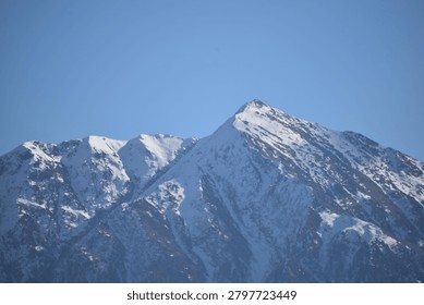 A sharp, pyramidal mountain peak rises prominently against a clear, solid blue sky. The steep slopes and jagged ridges of the summit are heavily blanketed with white snow and ice, emphasizing the deep vertical fissures and rocky contours of the mountain face.

To the left, additional craggy mountain peaks extend along the massive ridge line, also coated in patches of winter snow. Direct, bright daylight illuminates the right-facing slopes, casting the deep crevices and opposite aspects into crisp, dark shadows that contrast sharply with the sunlit snowy heights.