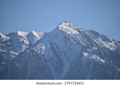 A sharp, pyramidal mountain peak rises prominently against a clear, solid blue sky. The steep slopes and jagged ridges of the summit are heavily blanketed with white snow and ice, emphasizing the deep vertical fissures and rocky contours of the mountain face.

To the left, additional craggy mountain peaks extend along the massive ridge line, also coated in patches of winter snow. Direct, bright daylight illuminates the right-facing slopes, casting the deep crevices and opposite aspects into crisp, dark shadows that contrast sharply with the sunlit snowy heights.