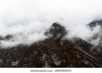 A sharp, rugged mountain peak pierces dramatically through a heavy shroud of rolling alpine mist in Yunnan Province, China. Dense white fog engulfs the lower ravines and sweeps across the craggy, heavily textured dark stone summit. The moody, high-contrast overcast lighting emphasizes the raw power, verticality, and mysterious isolation of high-altitude East Asian geological formations.