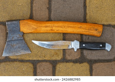 a sharp shiny dangerous old army iron knife with a black handle along with a small rusty hatchet lie on a concrete road during the day outdoors