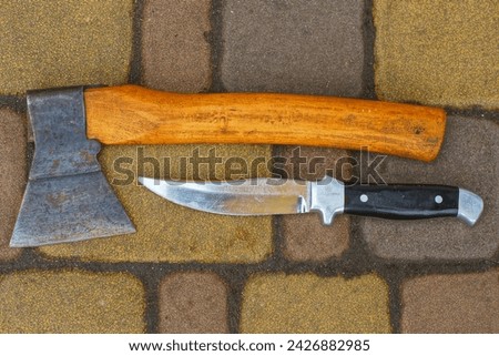 a sharp shiny dangerous old army iron knife with a black handle along with a small rusty hatchet lie on a concrete road during the day outdoors