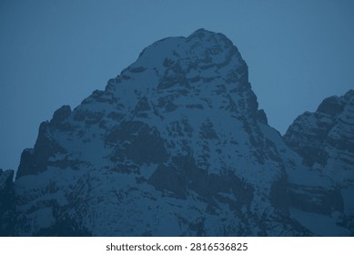 Sharp, snow-capped mountain peak rises against twilight blue sky. Rocky ridges gleam under soft, fading light. Majestic silence and calmness fill the high-altitude scene. Perfect for travel, adventure