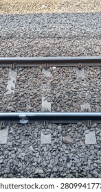 sharp top-down close-up photograph captures the gritty texture of heavy-duty steel railway tracks running horizontally across the frame. The composition showcases the solid concrete sleepers (railroad ties) secured tightly under the shiny metal rails, surrounded by a dense layer of crushed gravel ballast stones.