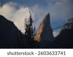 A sharp, towering peak rises high, resembling a pointed finger against the vast sky. Clouds swirl around its rugged edges, while sunlight highlights its dramatic slopes, creating a breathtaking sight.