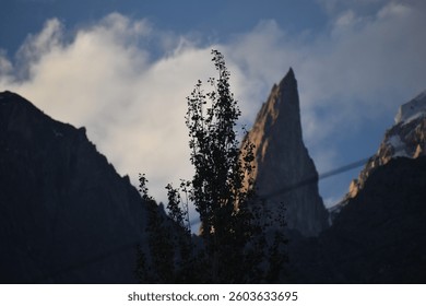 A sharp, towering peak rises high, resembling a pointed finger against the vast sky. Clouds swirl around its rugged edges, while sunlight highlights its dramatic slopes, creating a breathtaking sight.