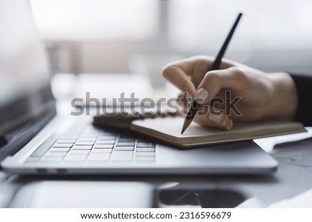 Sharp view of a woman's hand transcribing in a notepad on a cutting-edge laptop, with a defocused backdrop