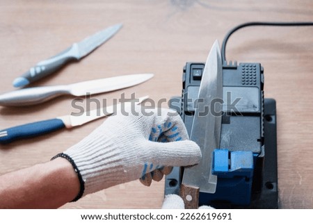 Sharpening a knife on an electric sharpener at home. The man's hand drives the knife blade between the blue sharpeners, dust flies on the machine.