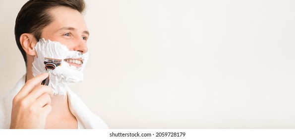 Shave, skin care, men's beauty banner with copy space for text. Side view of handsome smiling young man with moisturizing foam on face and towel on shoulders shaving modern sharp razor indoors.