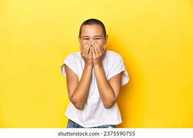 Shaved head woman in white tank top, yellow backdrop laughing about something, covering mouth with hands. - Powered by Shutterstock