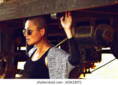 Shaved headed girl in black sun glasses and brown bronzed skin profile portrait on the vintage train and railway station scenery, fashion and beauty style model photo session with authentic background