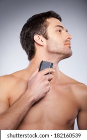 Shaving with electric shaver. Cropped image of handsome young man shaving his face with electric shaver while standing isolated on grey background