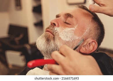 Shaving with a straight razor in a barbershop. A bearded old man being shaved in a barbershop. Classic shave by Stainless Steel Straight Edge Razor. - Shutterstock ID 2166571693