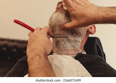 Shaving with a straight razor in a barbershop. A bearded old man being shaved in a barbershop. Classic shave by Stainless Steel Straight Edge Razor.