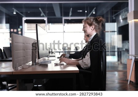She adjusts the cursor mid task, jacket slung over her shoulders, the beige divider and black desks melting into the moody dusk interior in a bright modern coworking office.