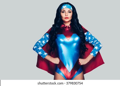 She can fight crime anywhere. Beautiful young woman in superhero costume looking at camera and keeping hands on hips while standing against grey background 