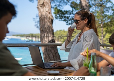 She drinks a cup of coffee while she works on laptop during summer vacation.A woman works on a summer vacation.