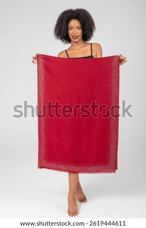 She lifts the ruby wrap in playful display; jeweled edges frame elegance while her bright smile and barefoot stance hint at carefree confidence and warm allure against a simple studio backdrop tonight