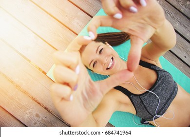 She loves sport. Top view of beautiful and young woman in sports clothing making heart with her hands and smiling while laying on yoga mat on wooden pier.