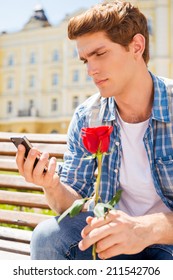 She Is Not Coming. Depressed Young Man Holding Single Rose And Looking At His Mobile Phone While Sitting On The Bench