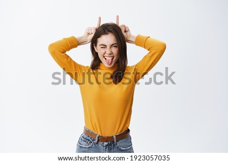 She is a rebel. Beautiful young woman showing tongue and finger devil horns on head, being stubborn, winking sassy at camera, standing against white background.