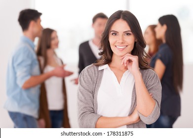 She is a team leader. Confident young woman holding hand on chin and smiling while group of people communicating on background