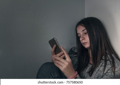  She Is A Victim Of Online  Social Networks. Sad Teen Checking Phone Sitting On The Floor In The Living Room At Home With A Dark Background. Victim Of Online Bullying Stalker Social Networks