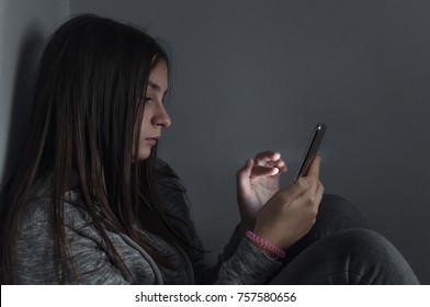  She Is A Victim Of Online  Social Networks. Sad Teen Checking Phone Sitting On The Floor In The Living Room At Home With A Dark Background. Victim Of Online Bullying Stalker Social Networks