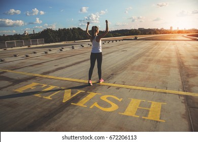She is a winner! Rear view of young and attractive woman in sports clothing keeping arms raised while passing finish line on the bridge with evening sunlight on background.