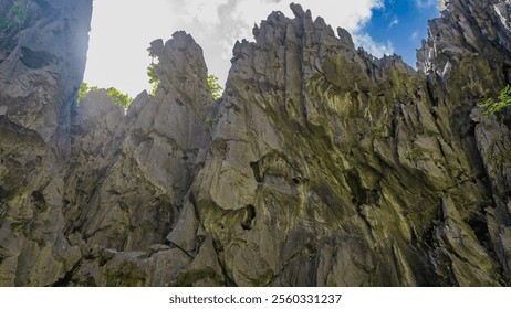 Sheer karst cliffs against a background of blue sky and clouds. Steep furrowed slopes, green vegetation, sharp peaks. Bottom-up view. Philippines. Palawan.