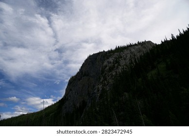 The Sheer Limestone Cliff: This dramatic perspective brings the rocky topography into sharp focus, showcasing a massive, near-vertical limestone cliff face. Weathered gray rock textures and deep vertical fractures dominate the center of the frame, illustrating the intense geological shearing and erosion typical of the Canadian Rockies.
