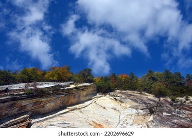 Sheer Rock Cliff Steeply Climbs To Tree Line.  Trees Are Turning Autumn Colors.  Sky Is Vivid Blue With Whispy Clouds.