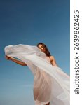A sheer veil billows dramatically in the wind as a woman holds it high against a bright blue sky. A minimalist visual of grace and movement.