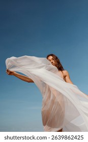 A sheer veil billows dramatically in the wind as a woman holds it high against a bright blue sky. A minimalist visual of grace and movement.