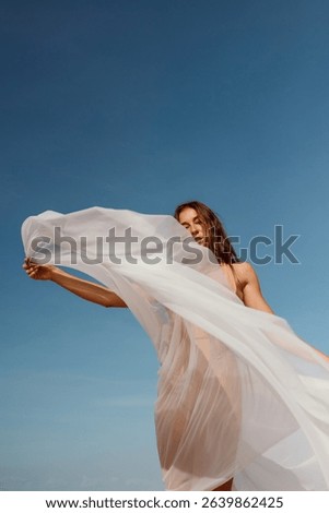 A sheer veil billows dramatically in the wind as a woman holds it high against a bright blue sky. A minimalist visual of grace and movement.