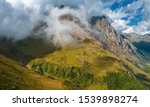 Sheer-sided rocky peak Kozhevnikov in fog rising up from the alpine meadows and river valley; rocky mountain ridge with steep stony slope; clouds coming down from tops; beautiful nature landscape
