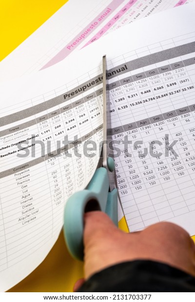 Sheet of the annual budget of any company with\
figures and concepts on a yellow background and blue scissors\
cutting the budget indicating the\
cuts.