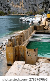 Sheet piling creates a dry work area for harbor development on a waterfront, with boats docked nearby and a dramatic rocky landscape beyond.