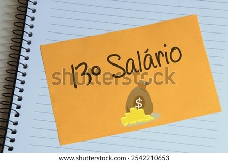Sheet of yellow post note paper or self-adhesive notes written 13º salary with background notebook. In portuguese 13º salário.