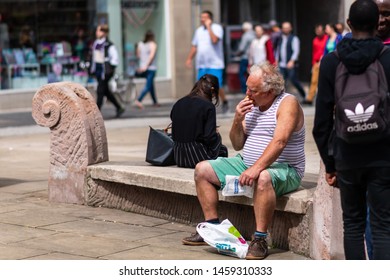 SHEFFIELD, UK - 20TH JULY 2019: An Elderly Overweight White Male In A Tanktop Sits On A Bench In The City Eating A Greggs Pasty