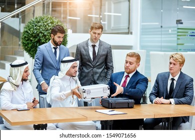 Sheikh In White Suit Give White Case With Money In Barter For Getting New Modern Technology By Caucasians With Black Case. Caucasian Men In Tuxedo. Sit On Table, Office Background