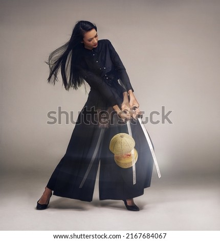 Shell slice you to pieces. Studio shot of a young woman in a martial arts outfit using a samurai sword to slice a melon in half.